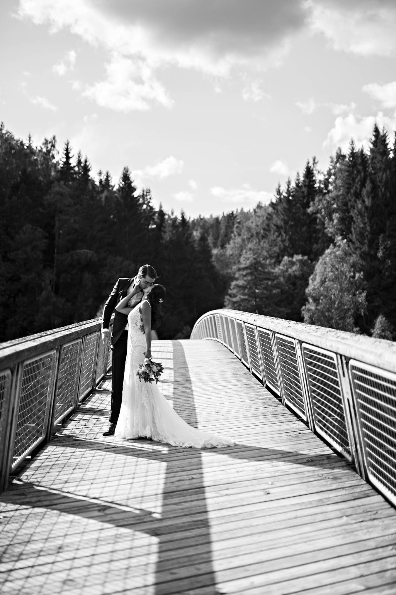 Brudepar på bro med skogen i bakgrunnen og fossen under. 