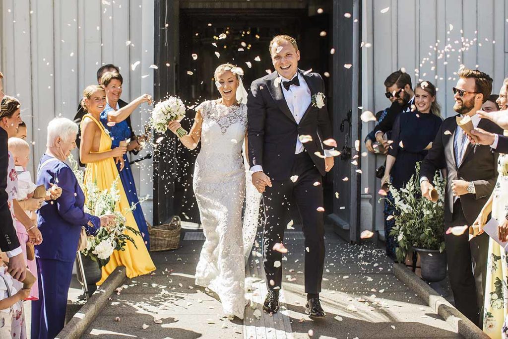 Nygift brudepar med blomster kastet på seg rett etter vielsen. Brud med hvit brudekjole og oppsatt hår. Brudebukett med peoner.