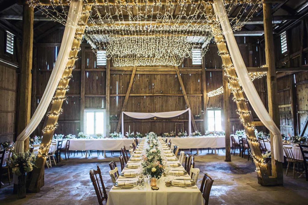 Låvebryllup i Drøbak med lysteppe, dekket bord og hvite gardiner. Blomstereng på bordene, dekketallerken i gull og gullbestikk.