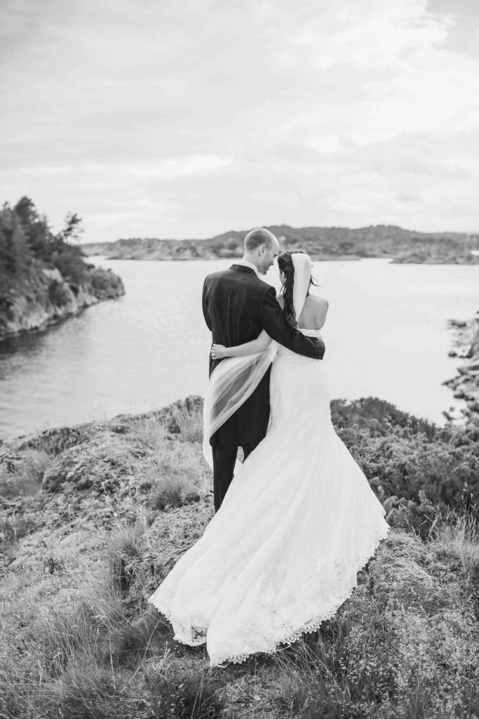 Brudepar på bryllupsdag, nyforelsket og smilende. Brud med hvit brudekjole og løst hår med utsikt over sjøen. Brud med lang brudekjole med hvitt slep.