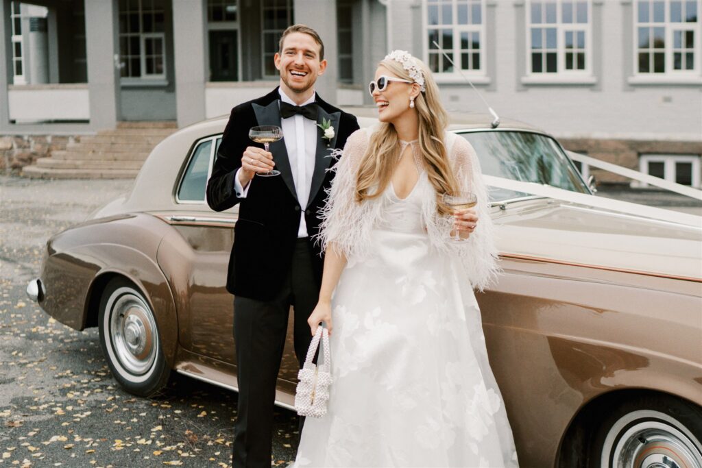 Et lykkelig, nygift par skåler med coupe champagneglass foran en veteranbil. De ser på hverandre og smiler kjærlig. Lys bakgrunn.