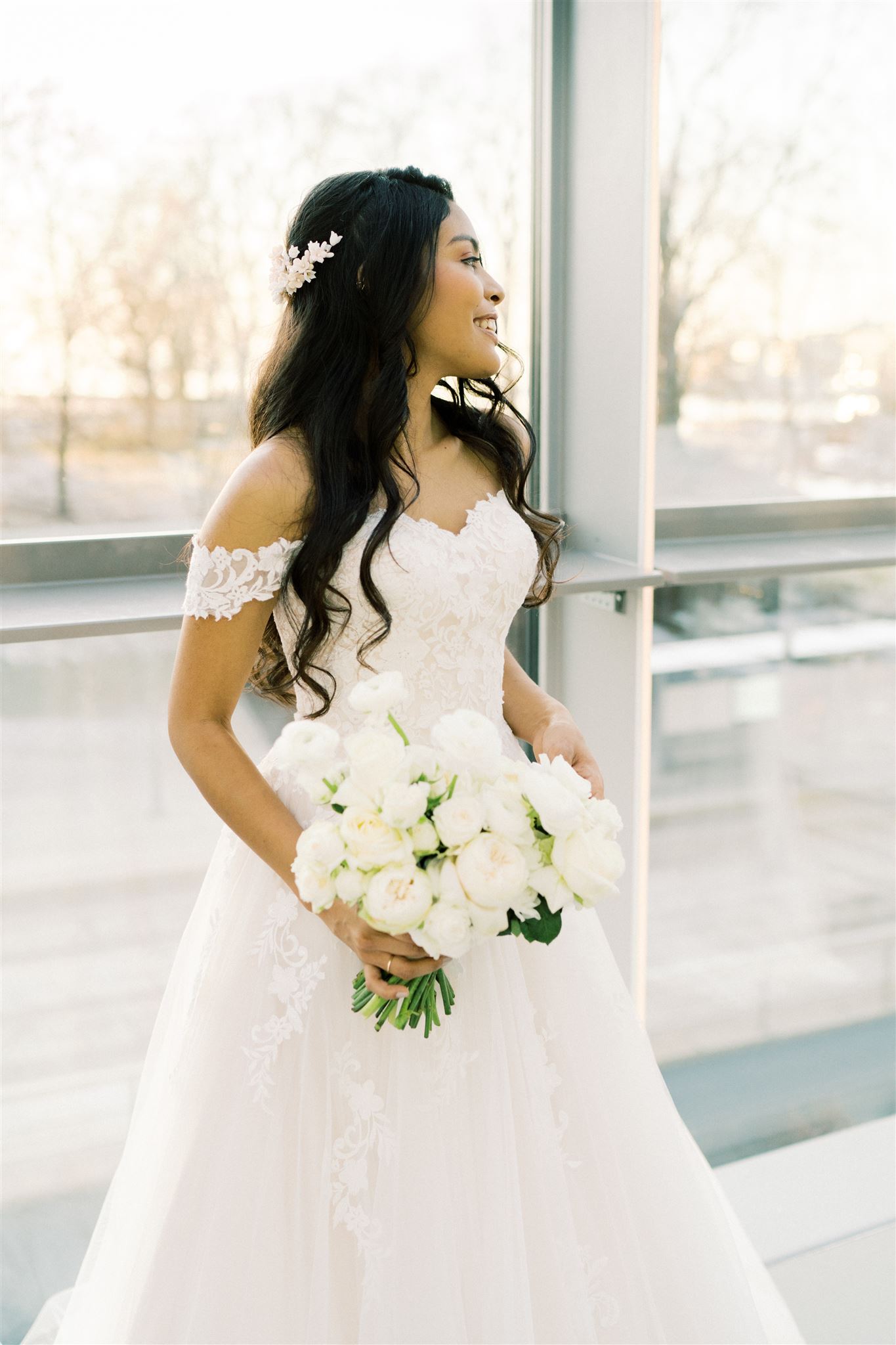 Vakker brud med løst krøllete hår og a-ilnjeformet brudekjole med blonder. Brudebuketten i hånden med vite blomster passer til hvit hodepynt i lyse blomsterfarger.
