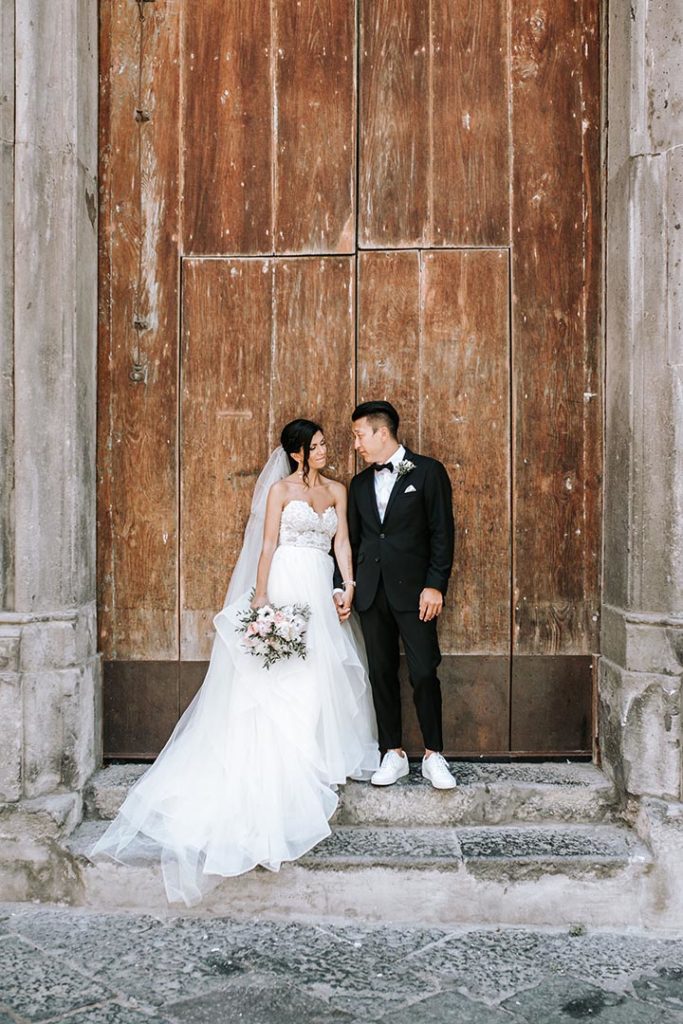 Brudepar i Sorrento foran en brun stor dør. Brud med brudebukett og hvit brudekjole med langt slep. Brudgom i smoking og hvite joggesko/sneakers.